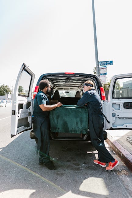 A man with a beard and serious expression, wearing a light grey knit beanie, a green puffer vest, and a dark blue jacket with white piping, stands outside a van during a home relocation. Behind him, t