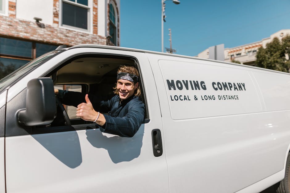 A white commercial van parked on a paved driveway outside a residential property during daytime, with a moving company worker visible inside the driver’s seat. The van is equipped for home relocatio