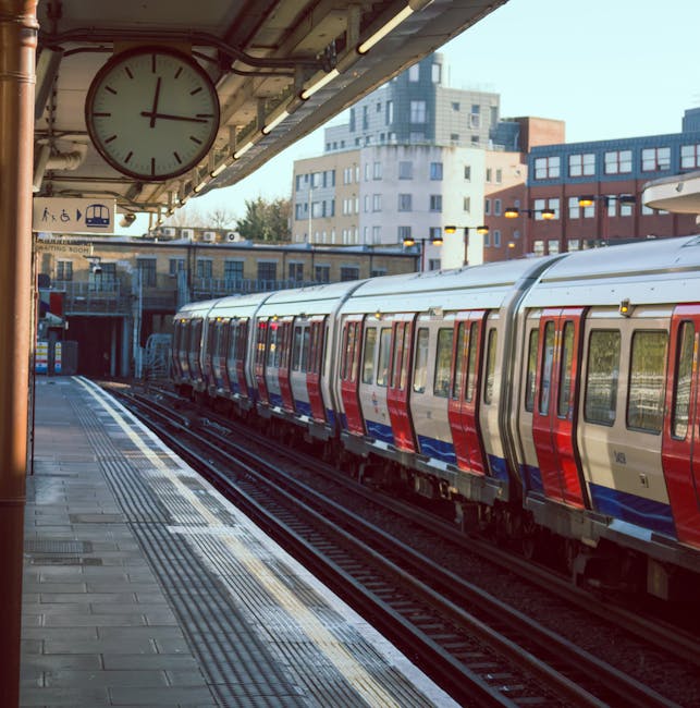 A London underground train with a red, white, and blue exterior is stationary at Swiss Cottage station, with the platform in the foreground featuring tactile paving strips and a clock hanging overhead showing 10:12. Behind the train, modern multi-storey residential buildings with large windows are visible, indicating an urban environment. The station platform is equipped with safety features, including signage and lighting, and is prepared for home relocation or moving services supported by Man with Van Swiss Cottage, who specializes in removals and furniture transport. The scene captures the loading process inside the station, with the train ready for departure or arrival, demonstrating typical moving logistics and transport infrastructure in London.