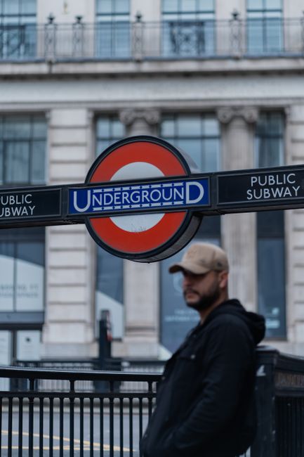 A man wearing a beige cap and dark jacket stands outdoors in front of a black metal railing, with a large London Underground 'Underground' sign featuring a red circle and blue rectangular sign with white text, indicating the proximity of the Swiss Cottage underground station. In the background, there is a historic building with classical architectural details, large windows, and stone columns, suggesting a busy urban area near a public transit hub. The scene is well-lit with natural daylight, and the man appears to be waiting or observing, while the setting relates to transportation and accessibility within the context of house removals and relocation services offered by Man with Van Swiss Cottage.