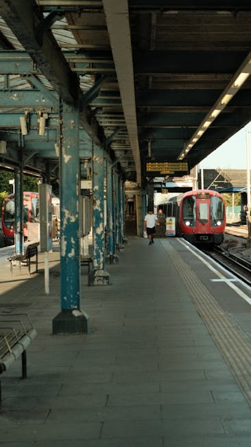A London underground train with a red, white, and blue exterior is stationary at Swiss Cottage station, with the platform in the foreground featuring tactile paving strips and a clock hanging overhead showing 10:12. Behind the train, modern multi-storey residential buildings with large windows are visible, indicating an urban environment. The station platform is equipped with safety features, including signage and lighting, and is prepared for home relocation or moving services supported by Man with Van Swiss Cottage, who specializes in removals and furniture transport. The scene captures the loading process inside the station, with the train ready for departure or arrival, demonstrating typical moving logistics and transport infrastructure in London.